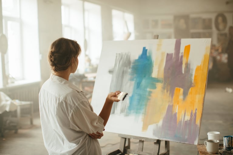 A female artist with a brush in her hands stands next to her artwork, thoughtfully gazing at it in a light, spacious studio