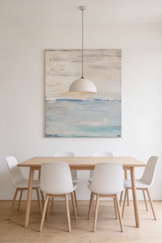 Minimalist dining room with wooden table, white chairs, pendant lamp, and abstract coastal artwork above