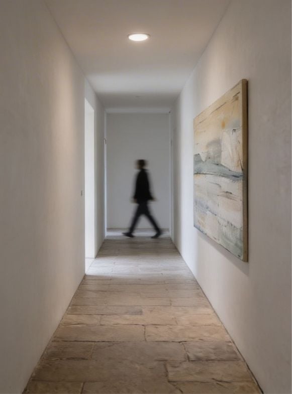 Blurred figure walking through a modern hallway with white walls, wooden flooring, framed artwork, and recessed ceiling light